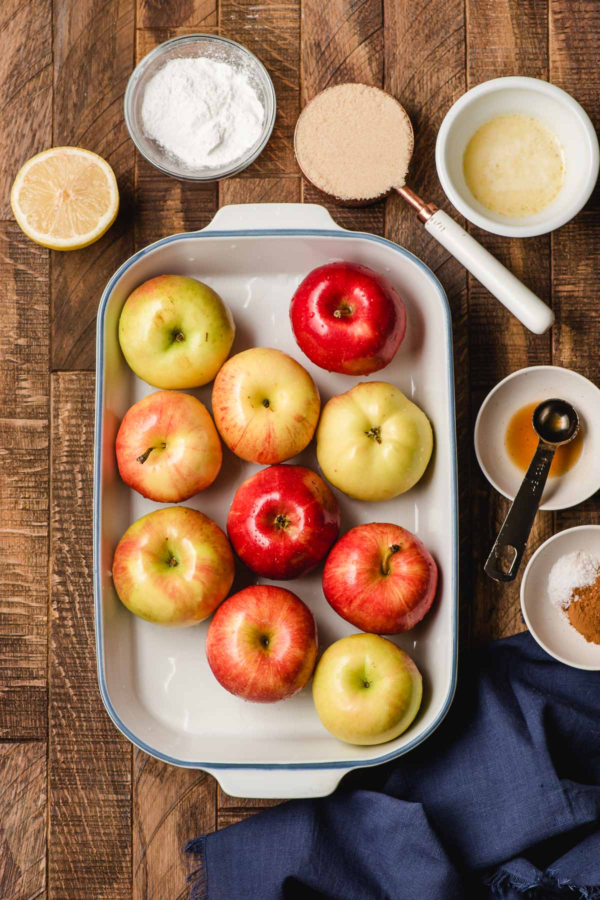 Whole apples in a casserole dish surrounded by bowls of brown sugar, melted butter, cornstarch, cinnamon, and a halved lemon.