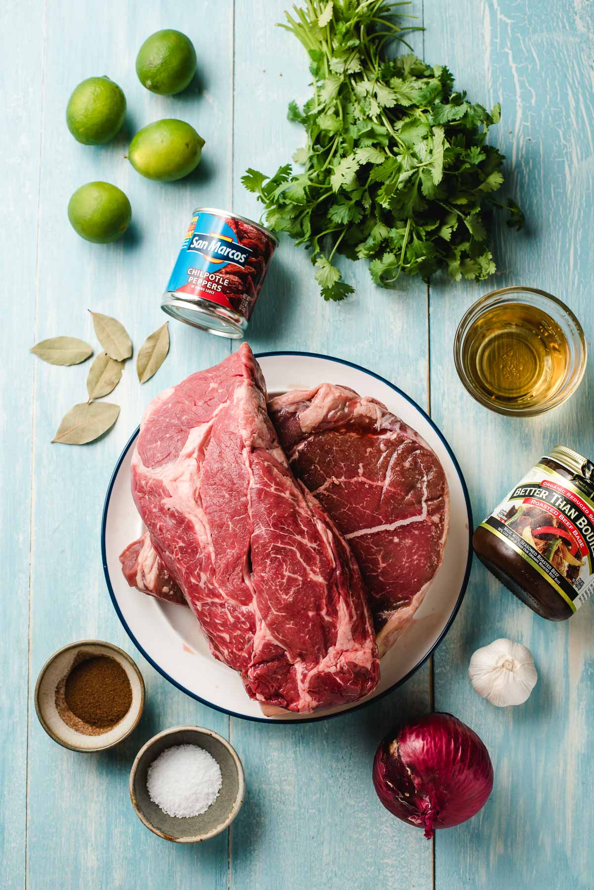 Ingredients for barbacoa beef on a light blue background- chuck roasts, salt, cumin and clove, lime, red onion, beef broth, cilantro, apple cider, chipotles in adobo.