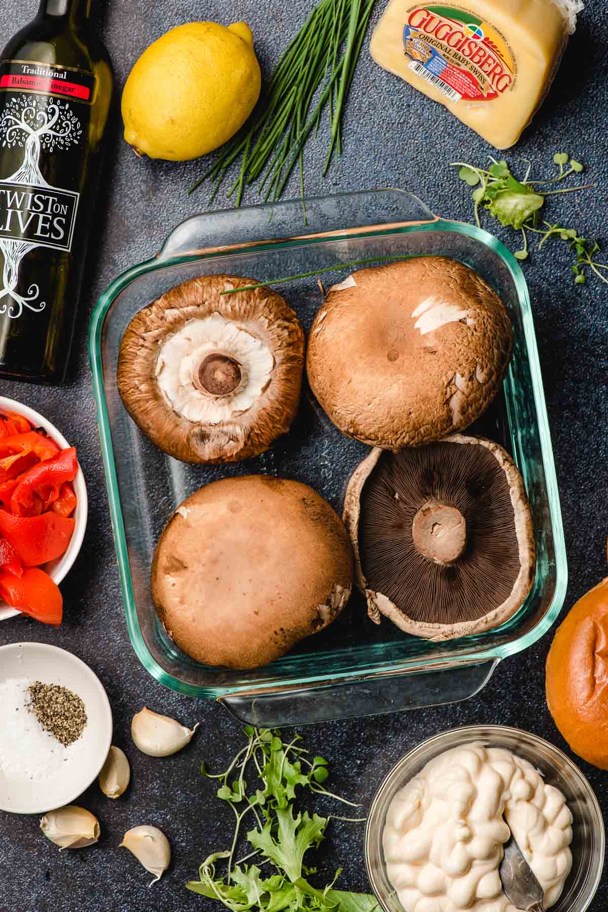 Portobello mushrooms in a glass casserole dish surrounded by ingredients for a sandwich.