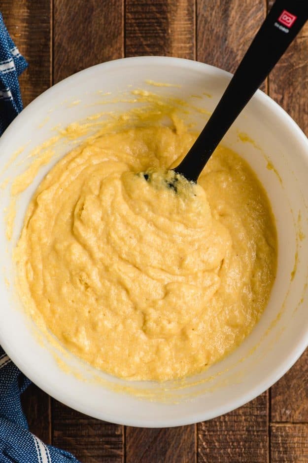 Cornbread batter in a white mixing bowl, being stirred by a black spatula.