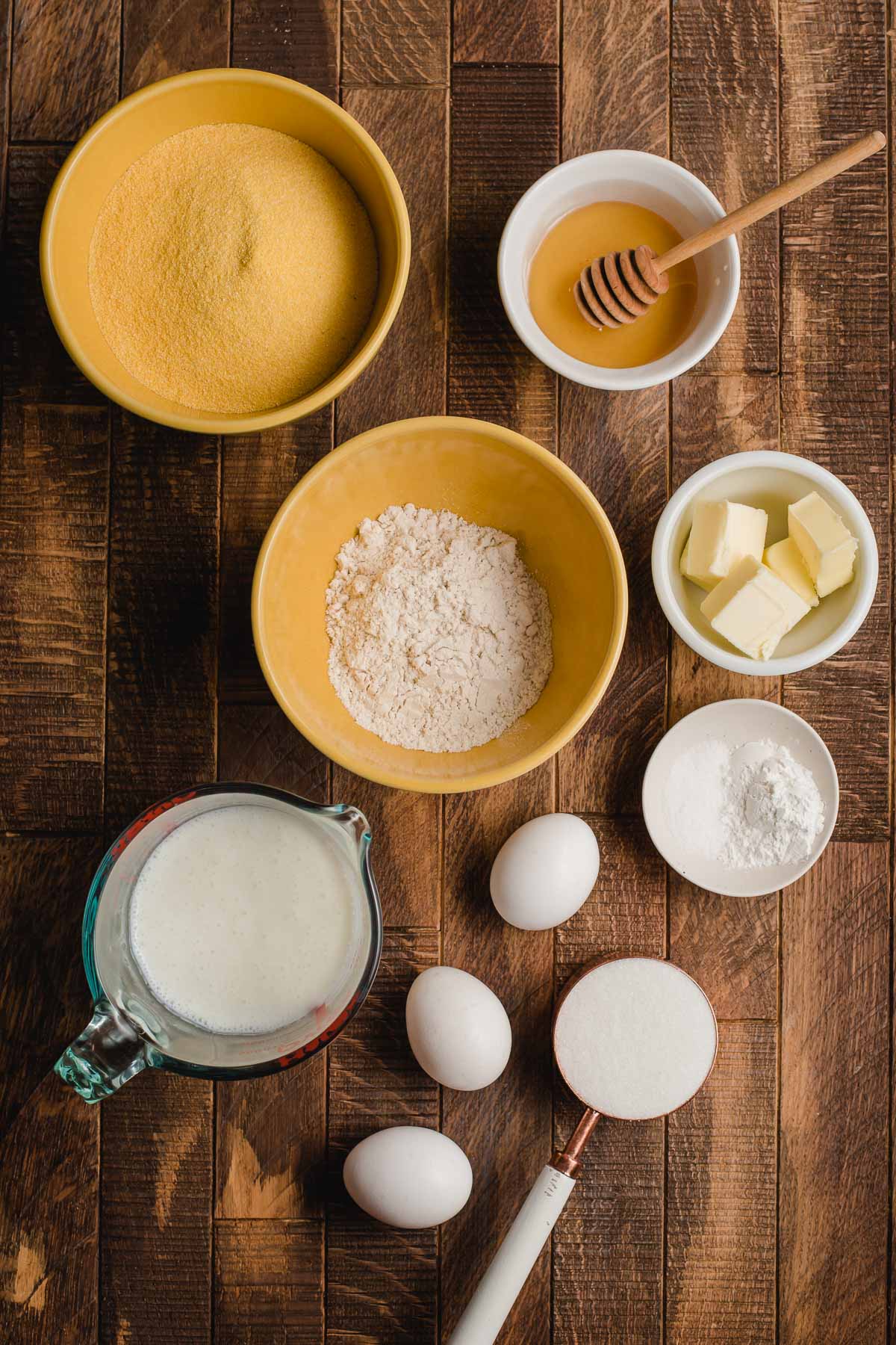 Ingredients on a wood surface--cornmeal, milk, eggs, sugar, butter, gluten free flour, honey, and baking soda.