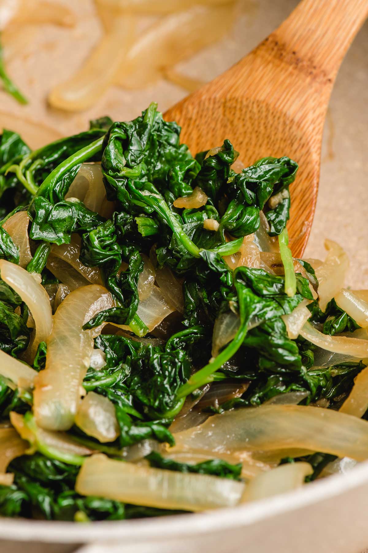 Wooden spoon scooping cooked spinach and onions out of a pan.