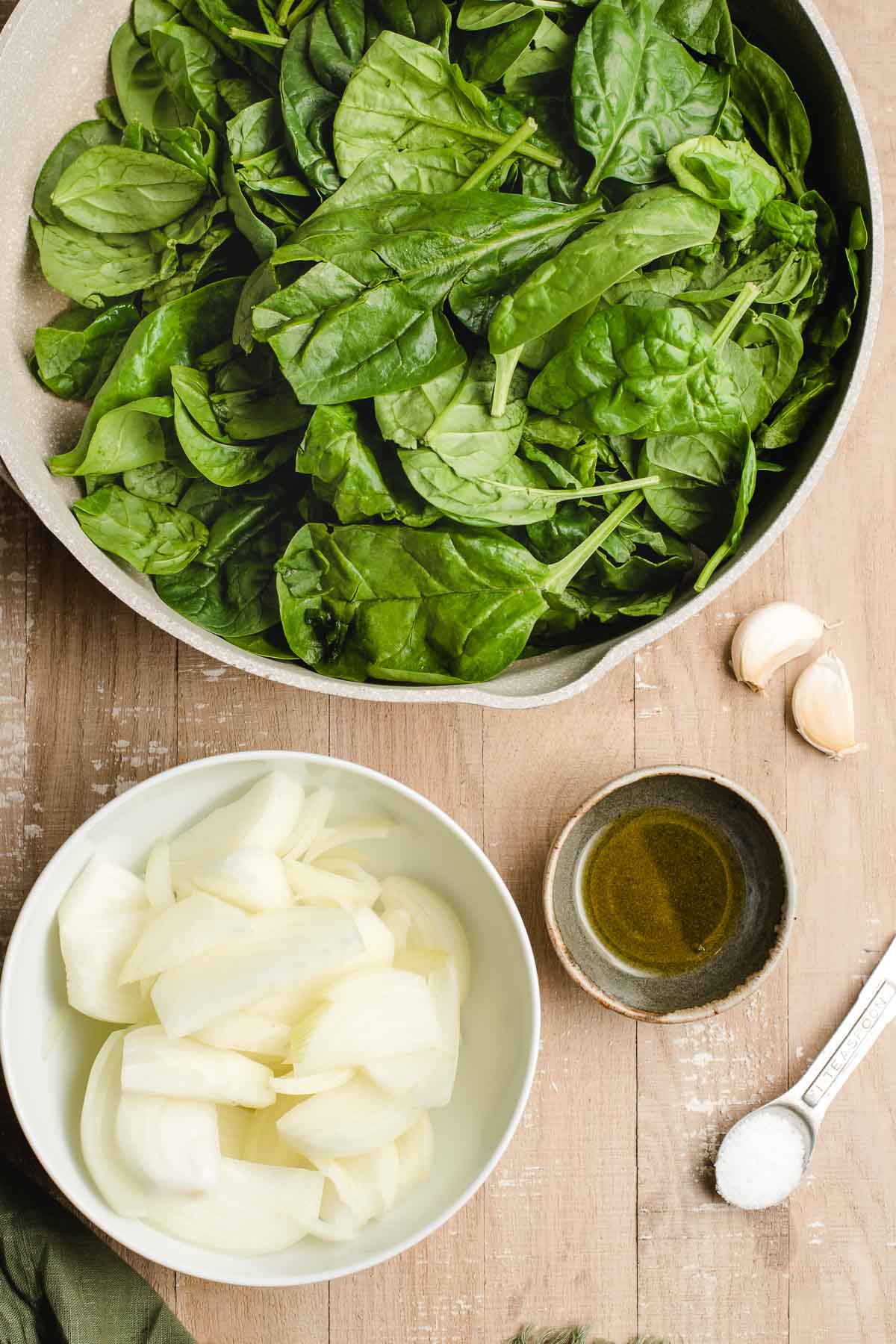 Skillet filled with raw spinach leaves, plu s a bowl of sliced onions, olive oil, salt, and garlic.