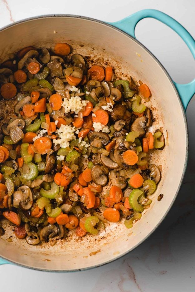 Dutch oven showing sauteed onions, carrots, celery, and mushrooms, with minced garlic on top.