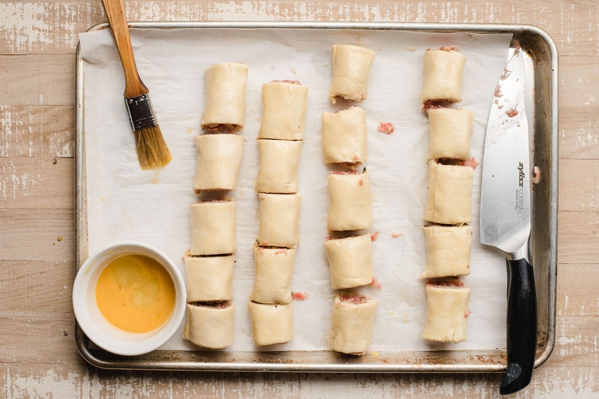 Unbaked sausage rolls on a baking sheet with a knife and egg wash.