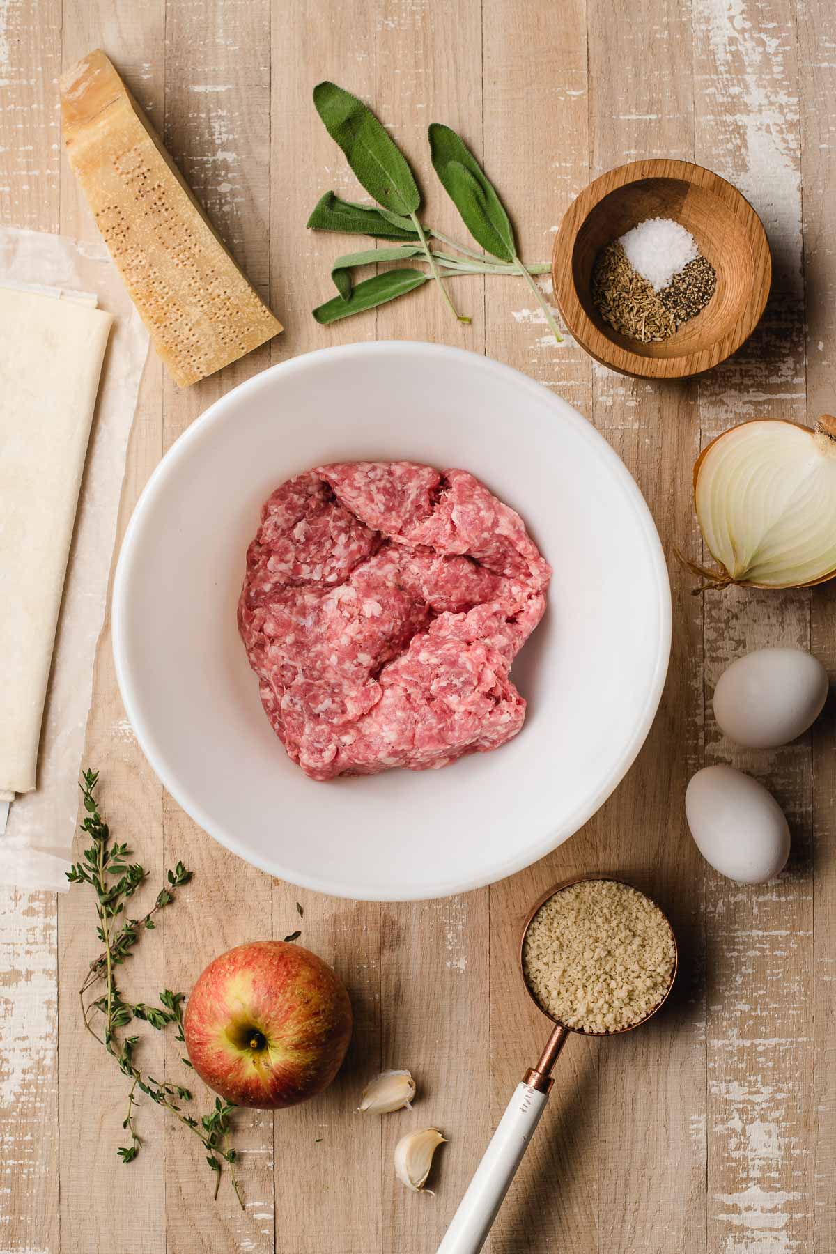 An array of ingredients on a wood background: ground pork, apple, breadcrumbs, eggs, ohions, sage, thyme, Parmesan cheese wedge, and puff pastry.