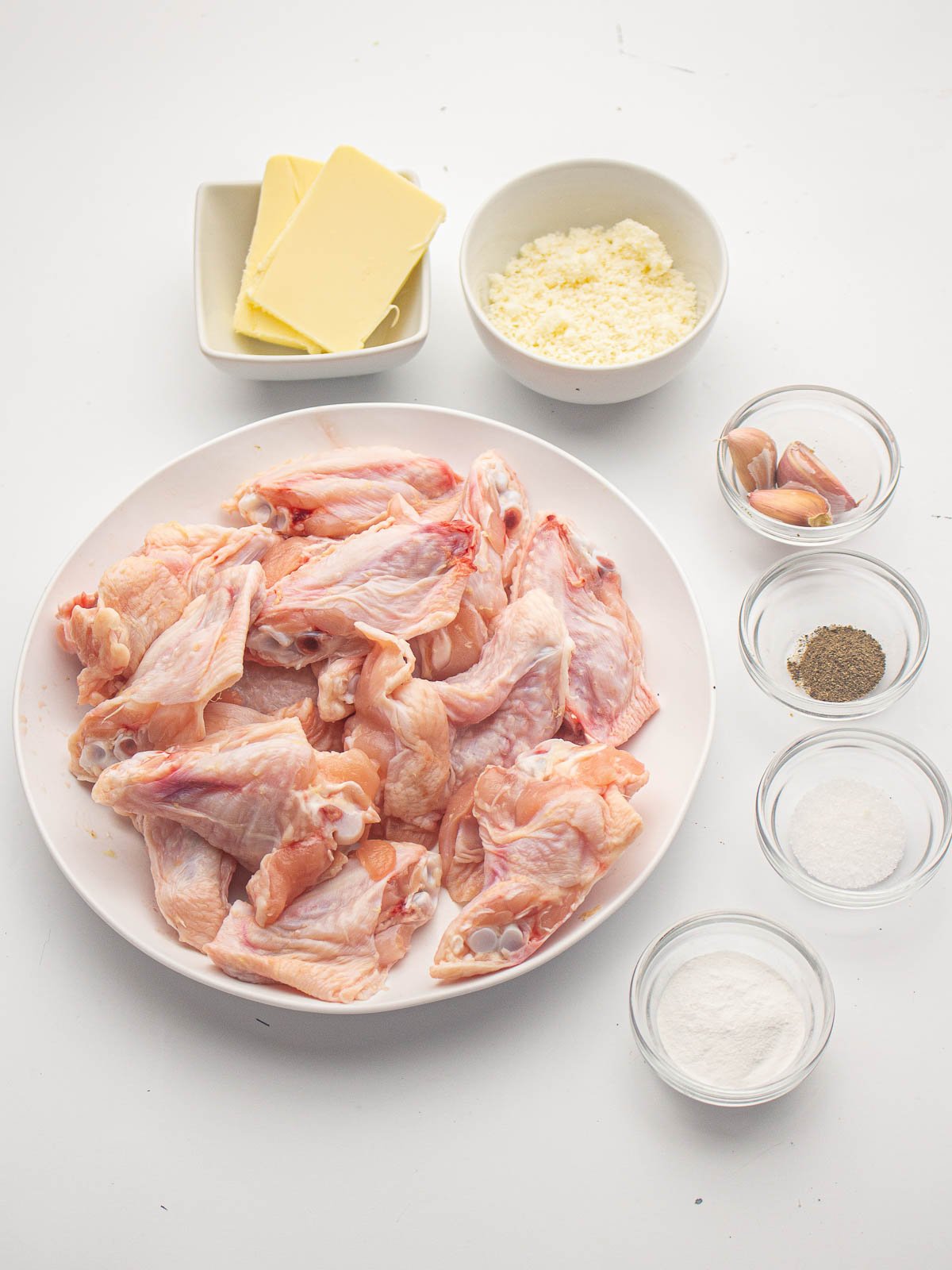 Ingredients for garlic parmesan wings--raw wings on a white plate and bowls of butter, grated Parmesan cheese, garlic, salt, pepper, and baking powder.