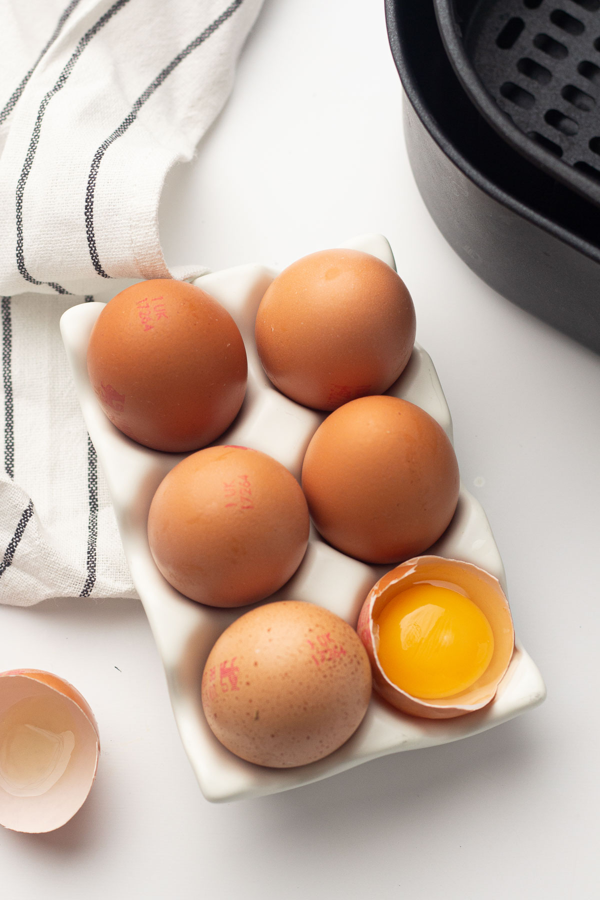 White egg tray holding six brown eggs. One egg is cracked in half to show the yolk inside.