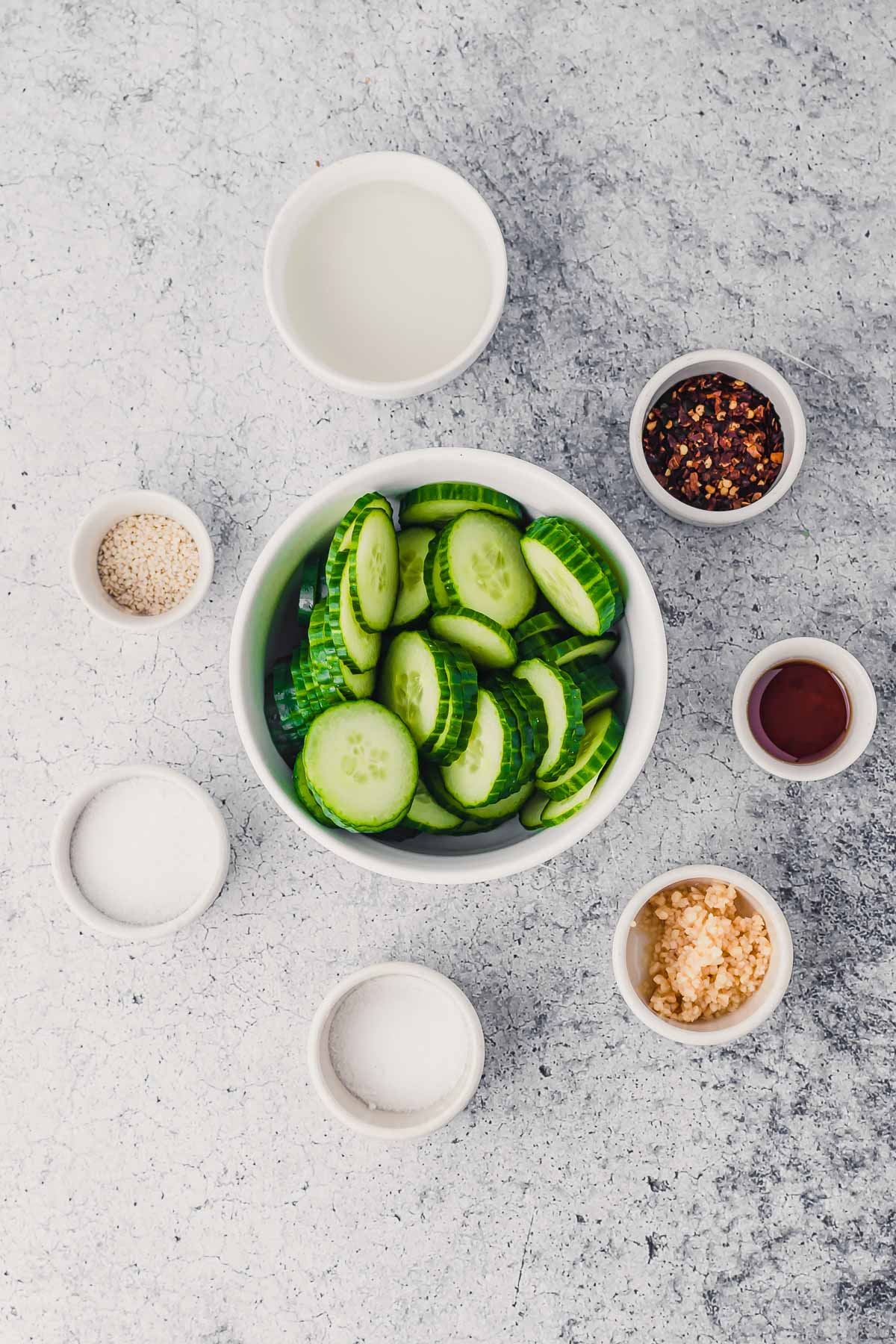 Ingredient to make Korean Pickled Cucumbers shown in bowls: sliced cucumbers, sugar, chili flakes, garlic, salt, rice vinegar, sesame oil.