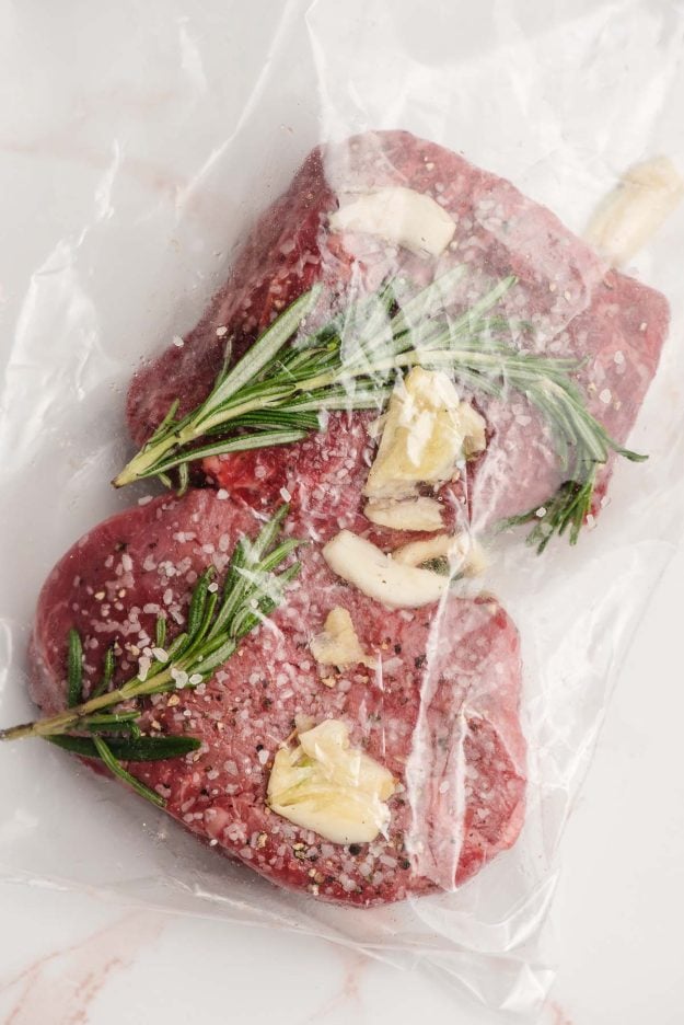Two filet mignon steaks in a bag with rosemary and garlic.