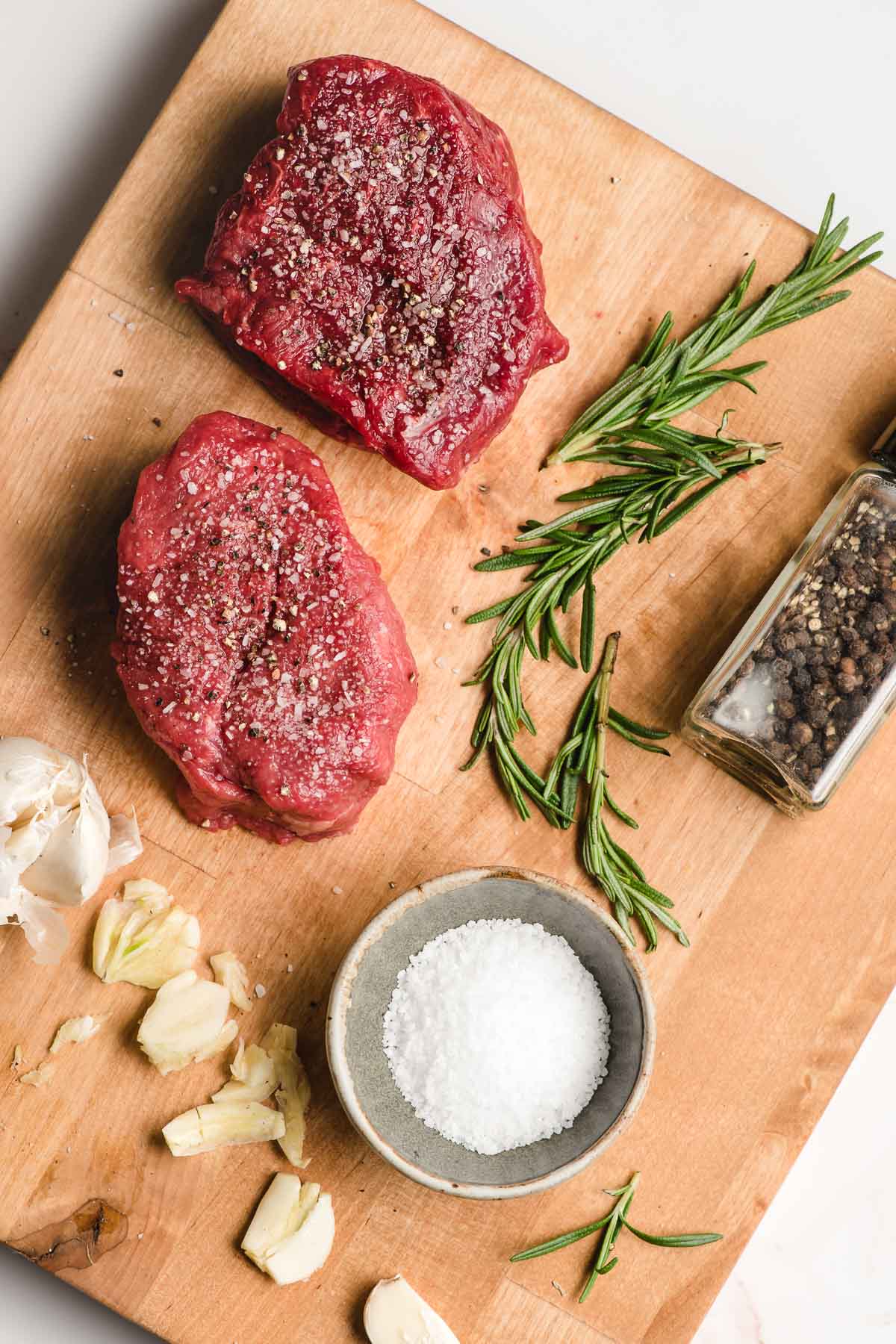 Two filet mignon steaks on a cutting board along with smashed garlic, salt, and rosemary sprigs.