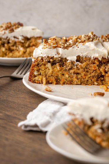Frosted round carrot cake shown with a slice taken out of it on a white cake platter.
