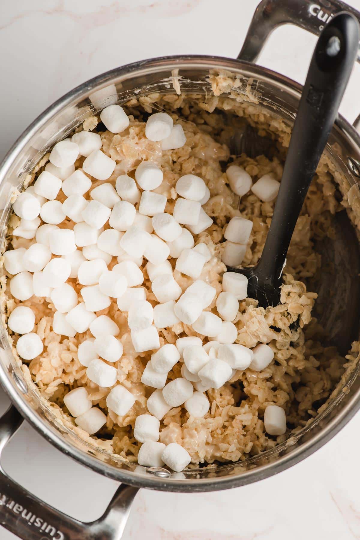 Mini marshmallows sprinkled over rice krispies in a big pot.