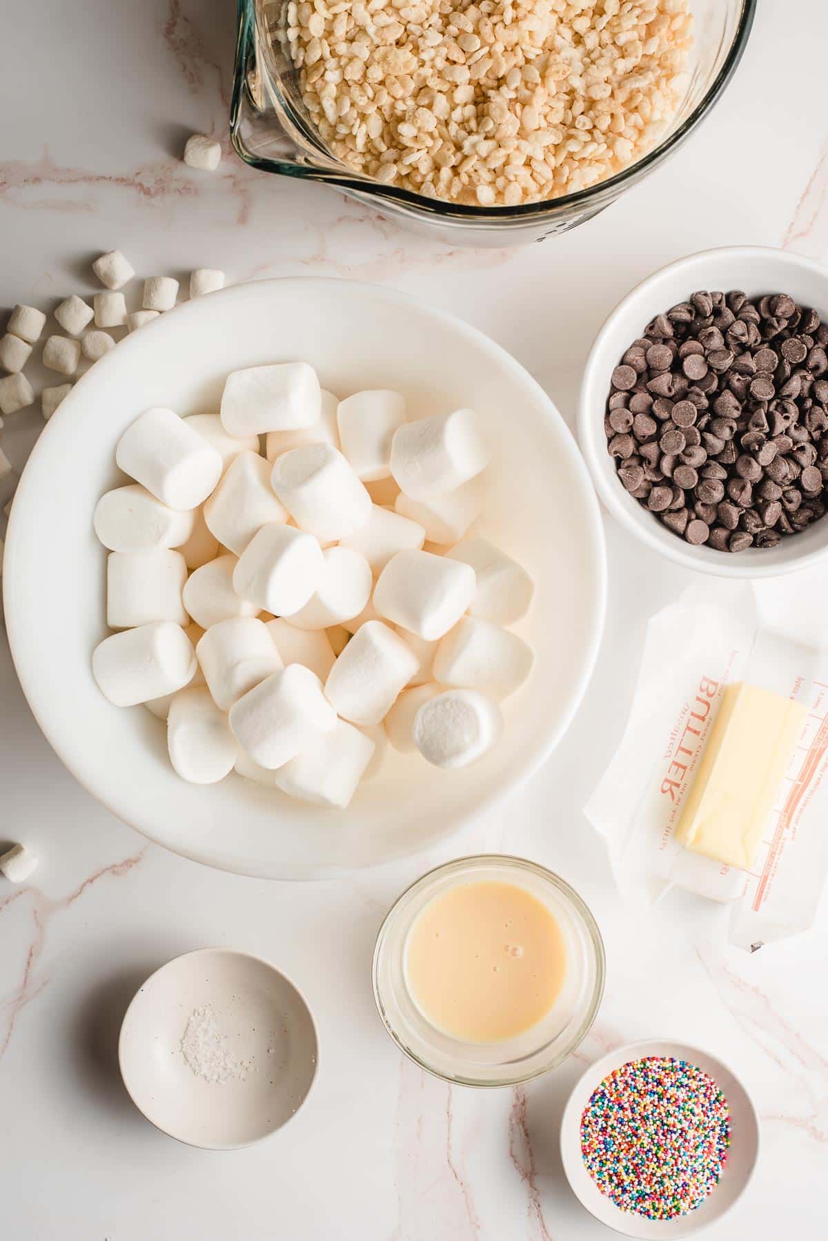 Ingredients in bowls-- marshmallows, butter, rice krispies, sweetened condensed milk, salt, sprinkles, and chocolate.