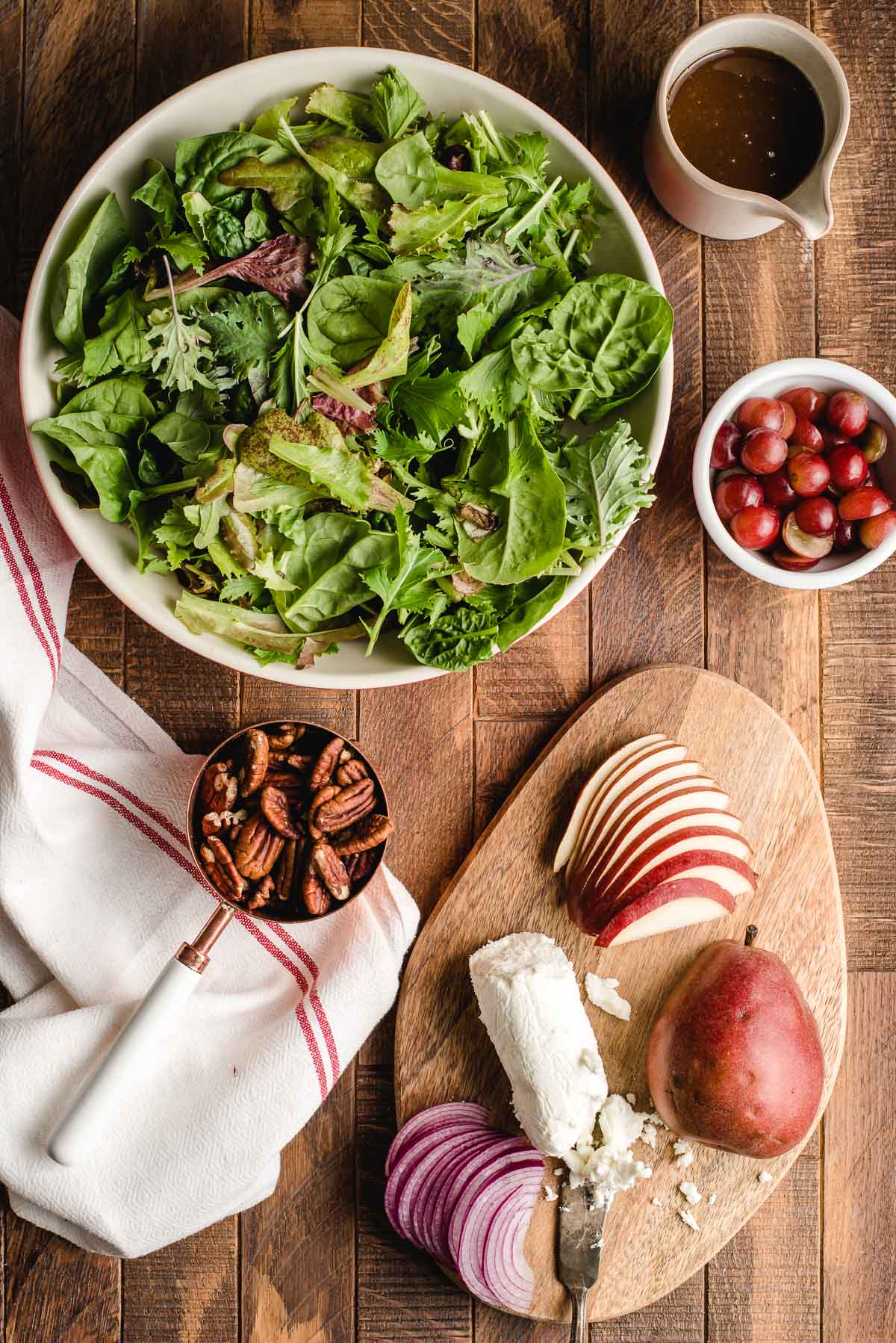 Bowl of mixed greens with bowls of pecans, cranberries, sliced pearsw, goat cheese, and maple dressing.