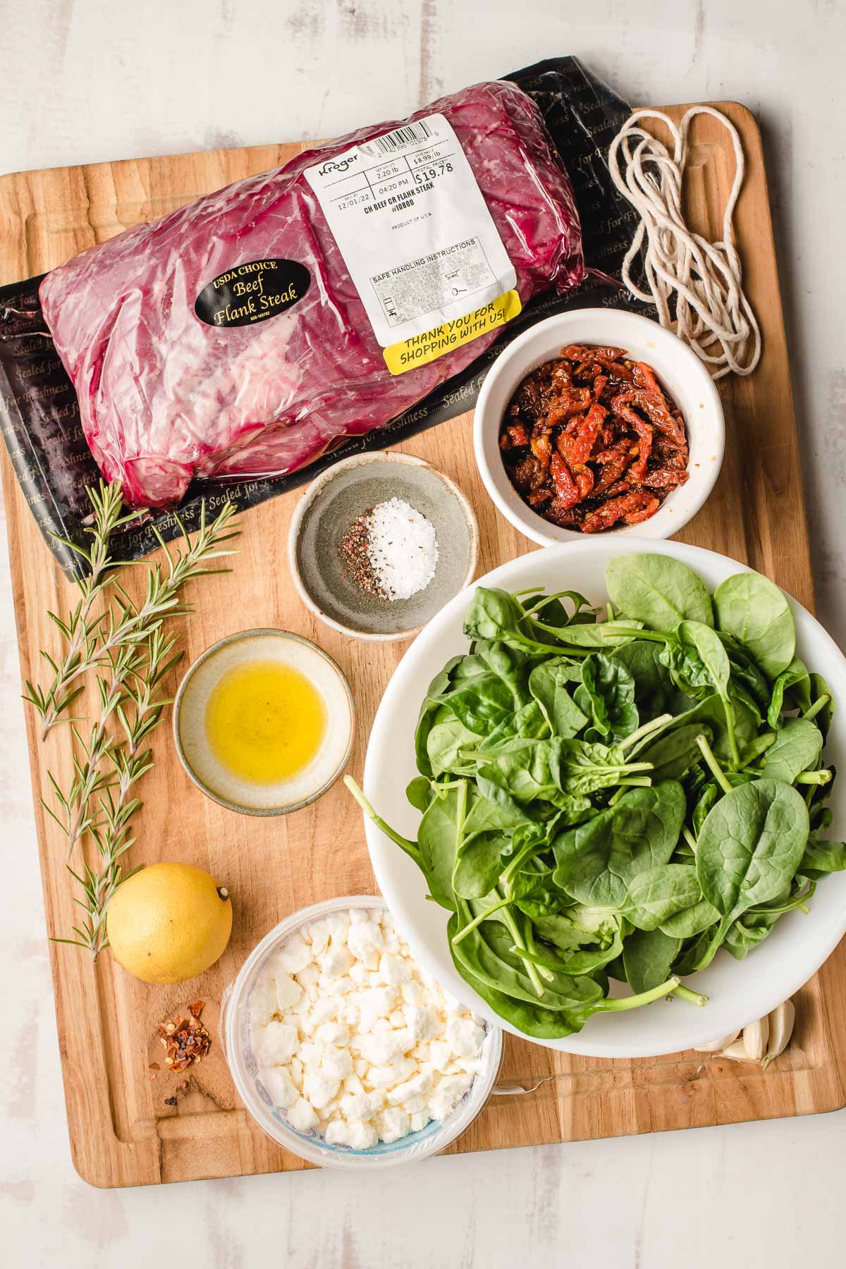 Cutting board with a package of flank steak, fresh spinach, olive oil, lemon, salt, sun dried tomatoes, and feta.