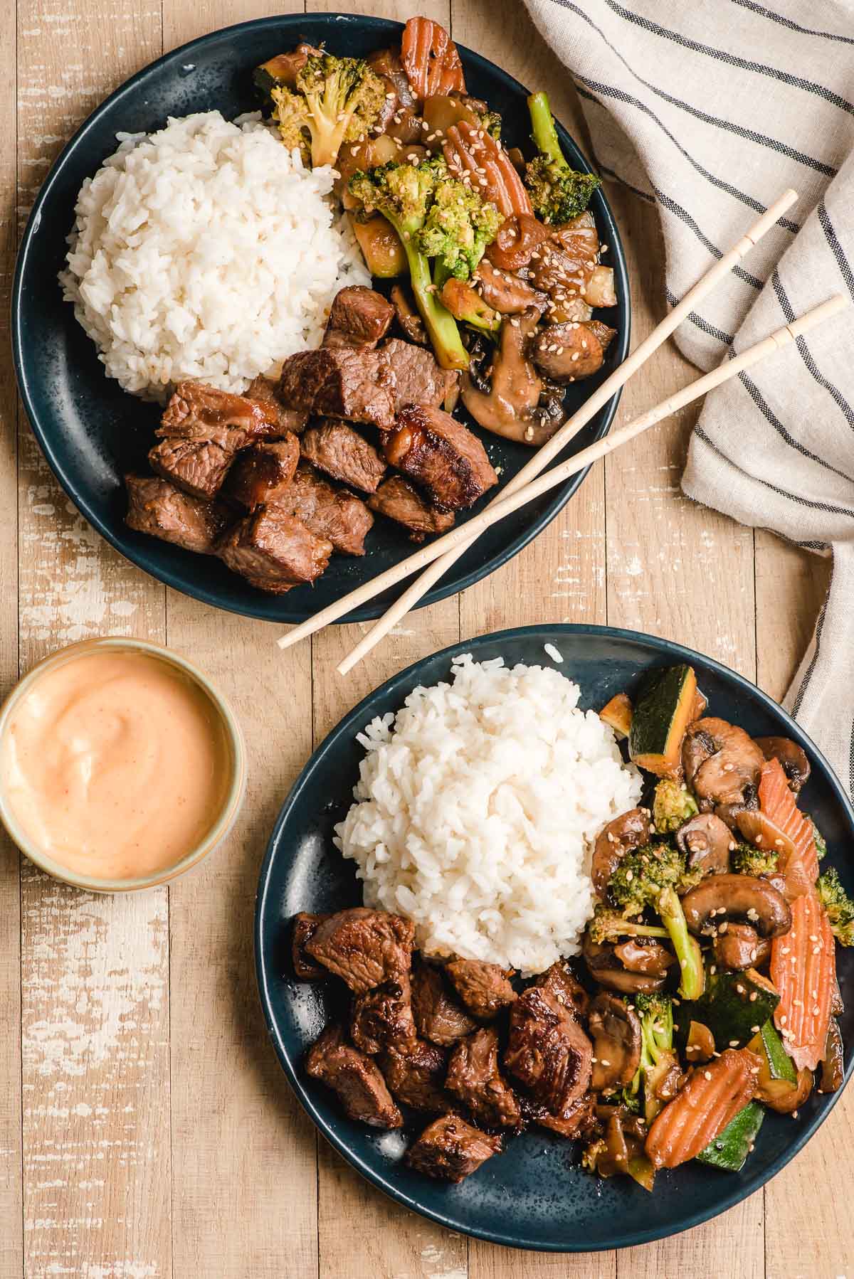 Two blue plates, each with Japanese Steakhouse steak bites, white rice, and hibachi veggies, plus a bowl with yum yum sauce.