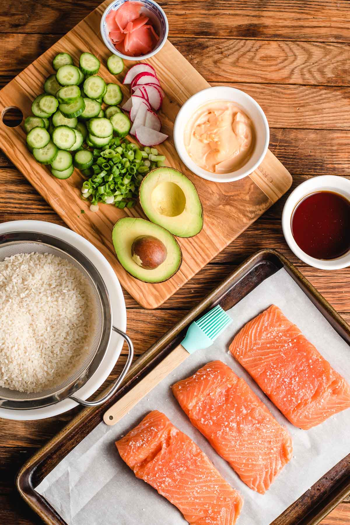 Salmon fillets on a sheet pan, rice in a colander, and cucumber, sliced avocado, and bowls of spicy mayo and eel sauce on a cutting board.