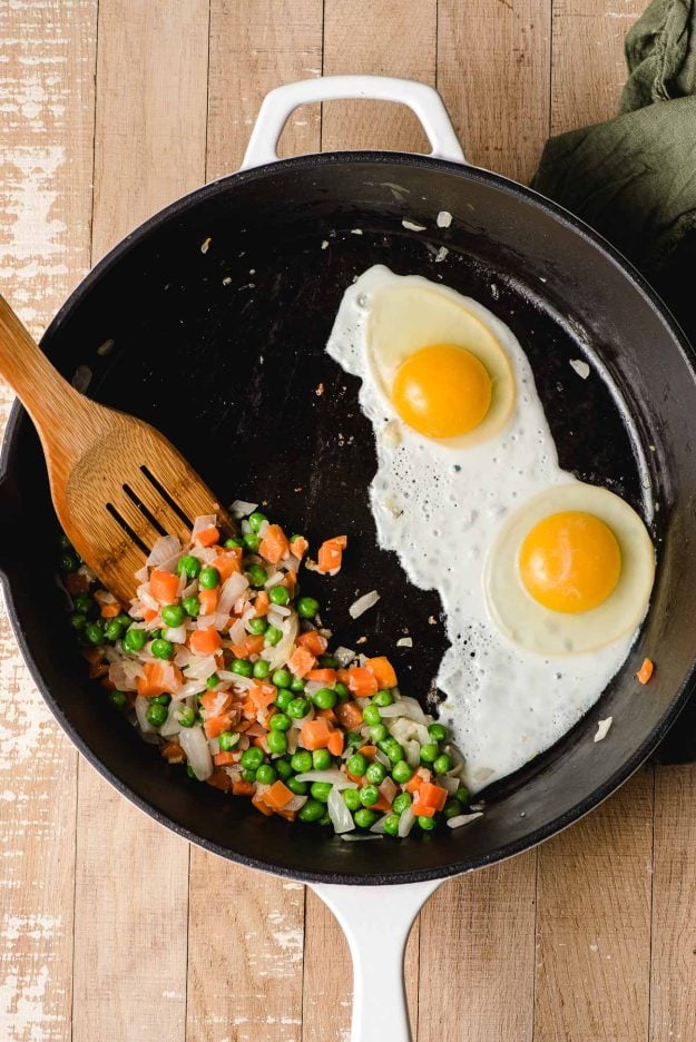 Two eggs frying in a pan next to sauteed onions, peas, and carrot.s
