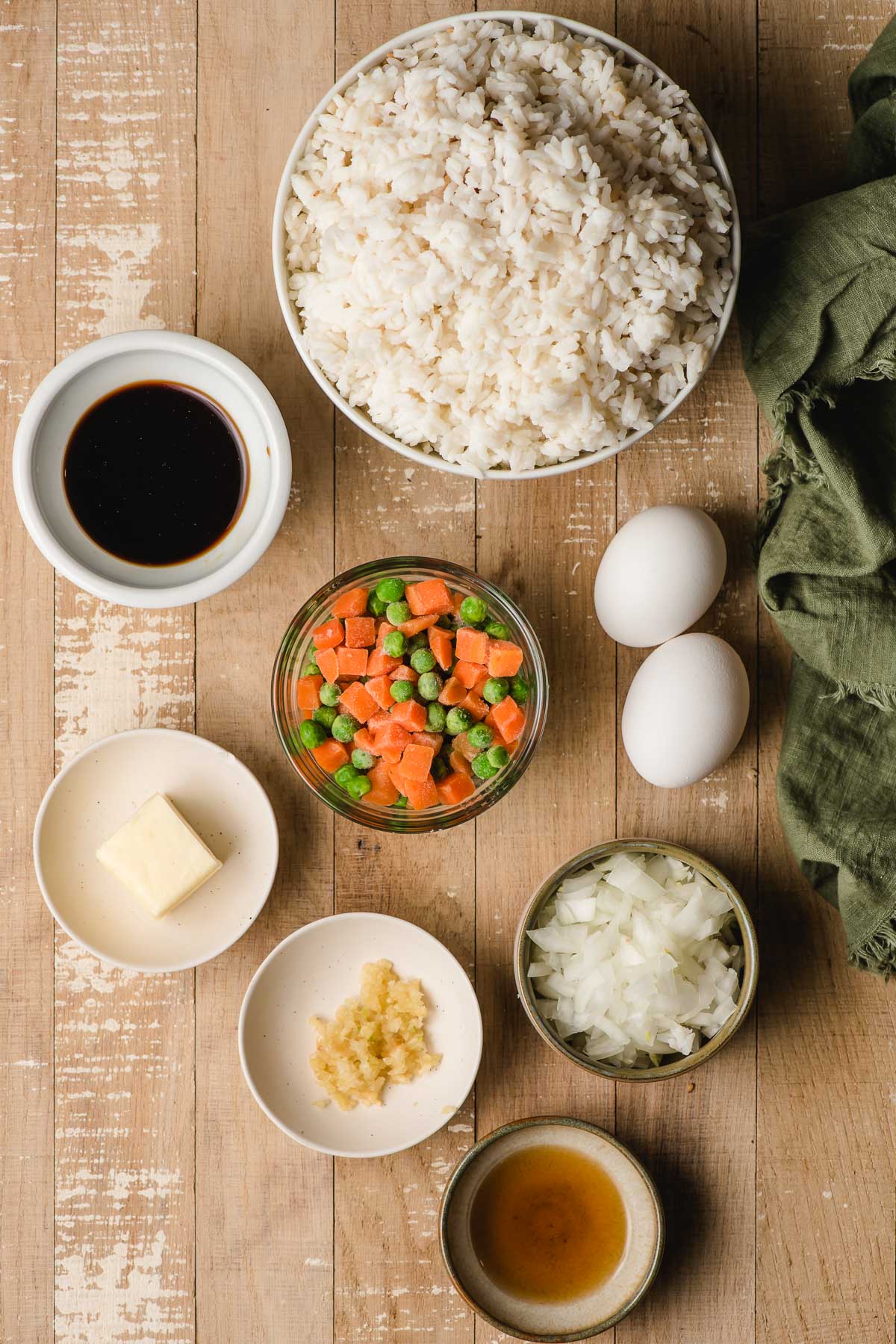 Several bowls filled with white rice, peas and carrots, butter, garlic, soy sauce, onion, and eggs.