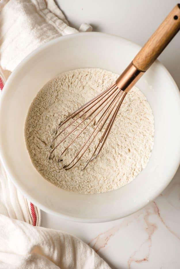 Flour, salt, and baking powder in a white mixing bowl with gold whisk.
