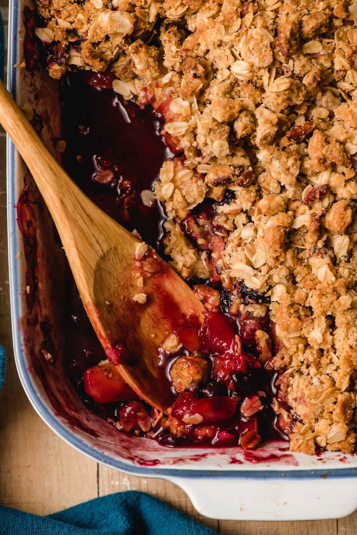 Rich, purple hued blueberry and apple crumble shown in a baking dish with a wooden spoon.