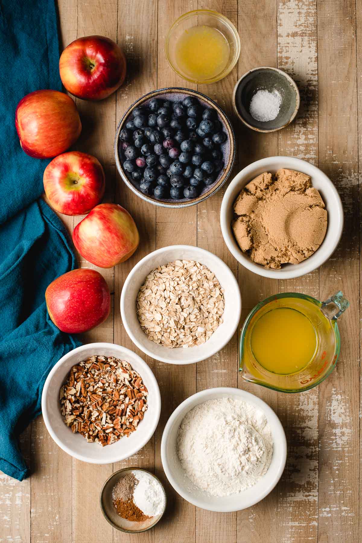 Apples, blueberries, brown sugar, oats, pecans, flour, butter, cinnamon, cardamom, and lemon displayed in bowls on a wood background.