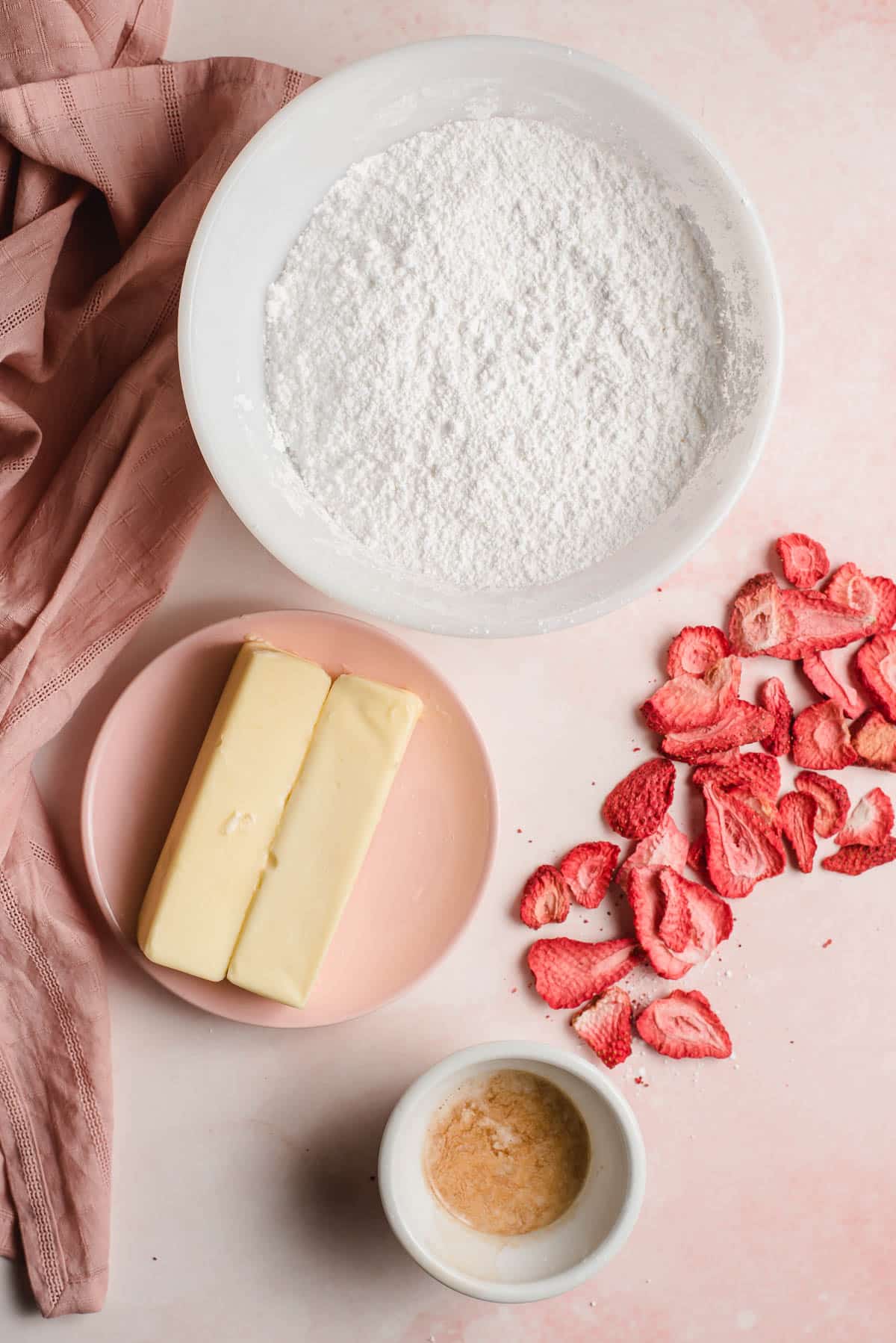 Powdered sugar, butter, zestless strawberries, and vanilla pericope shown in bowls on a pink background.