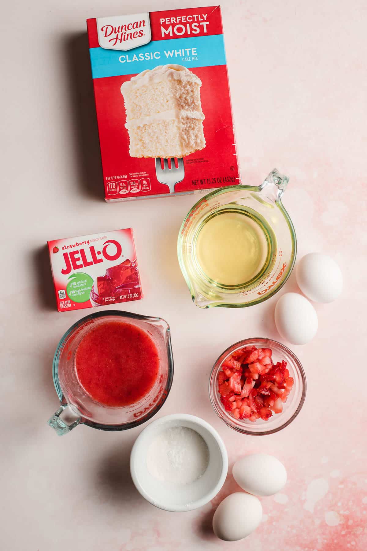 White confection mix, strawberry jello, pureed strawberries, eggs, oil, flour, and diced strawberries on a pink background.