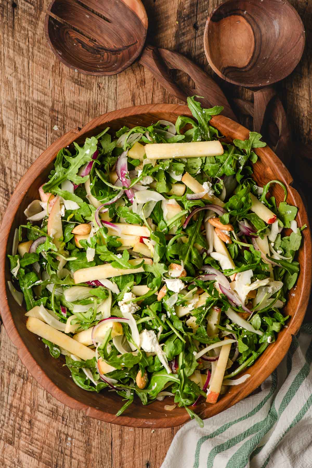 Arugula salad with fennel, apple, goat cheese, and red onion in a wooden trencher with decorative wooden salad spoons.