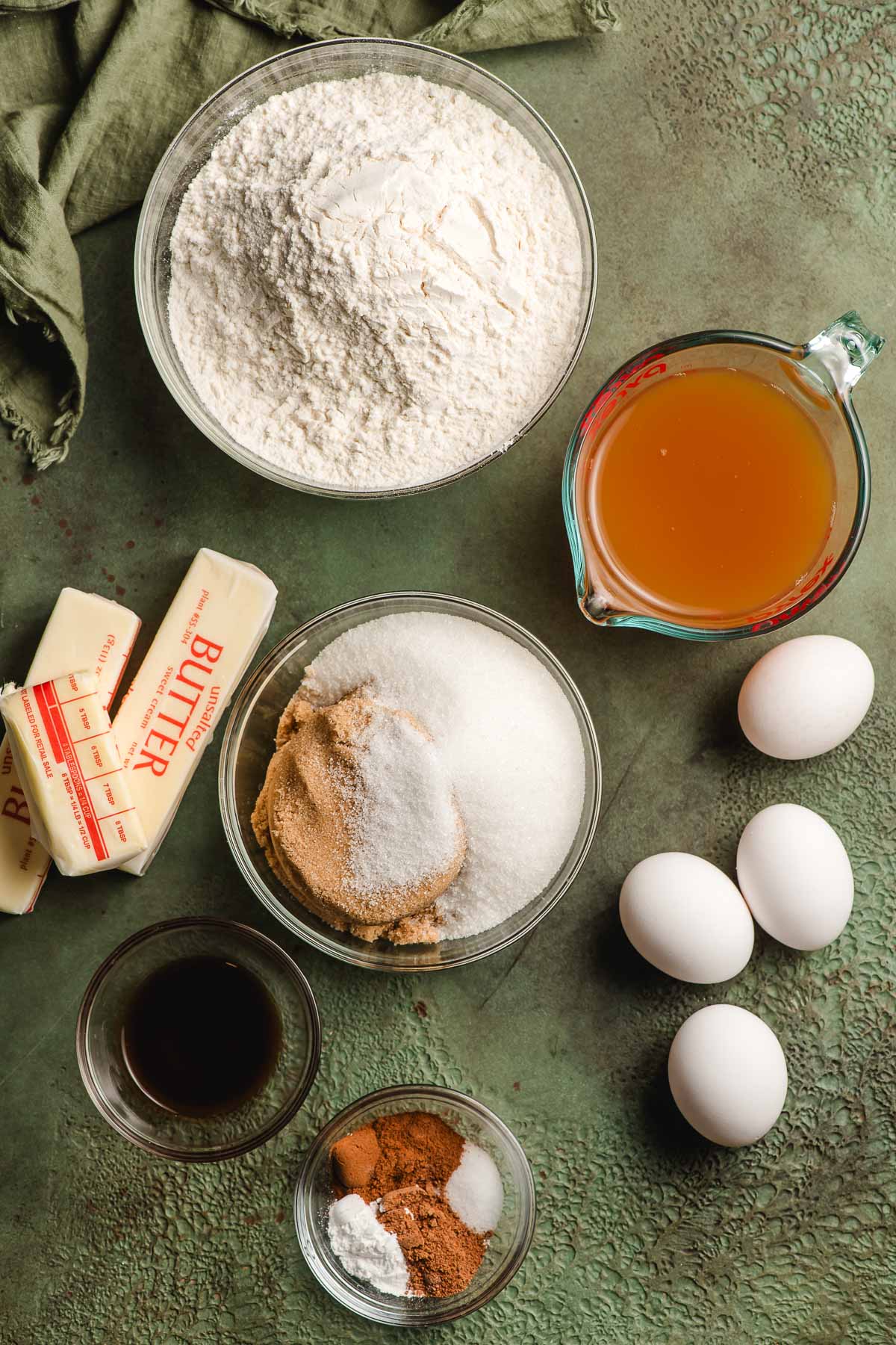 Flour, baking powder, cinnamon, nutmeg, salt, eggs, vanilla, butter, and apple cider in prep bowls on a green background.
