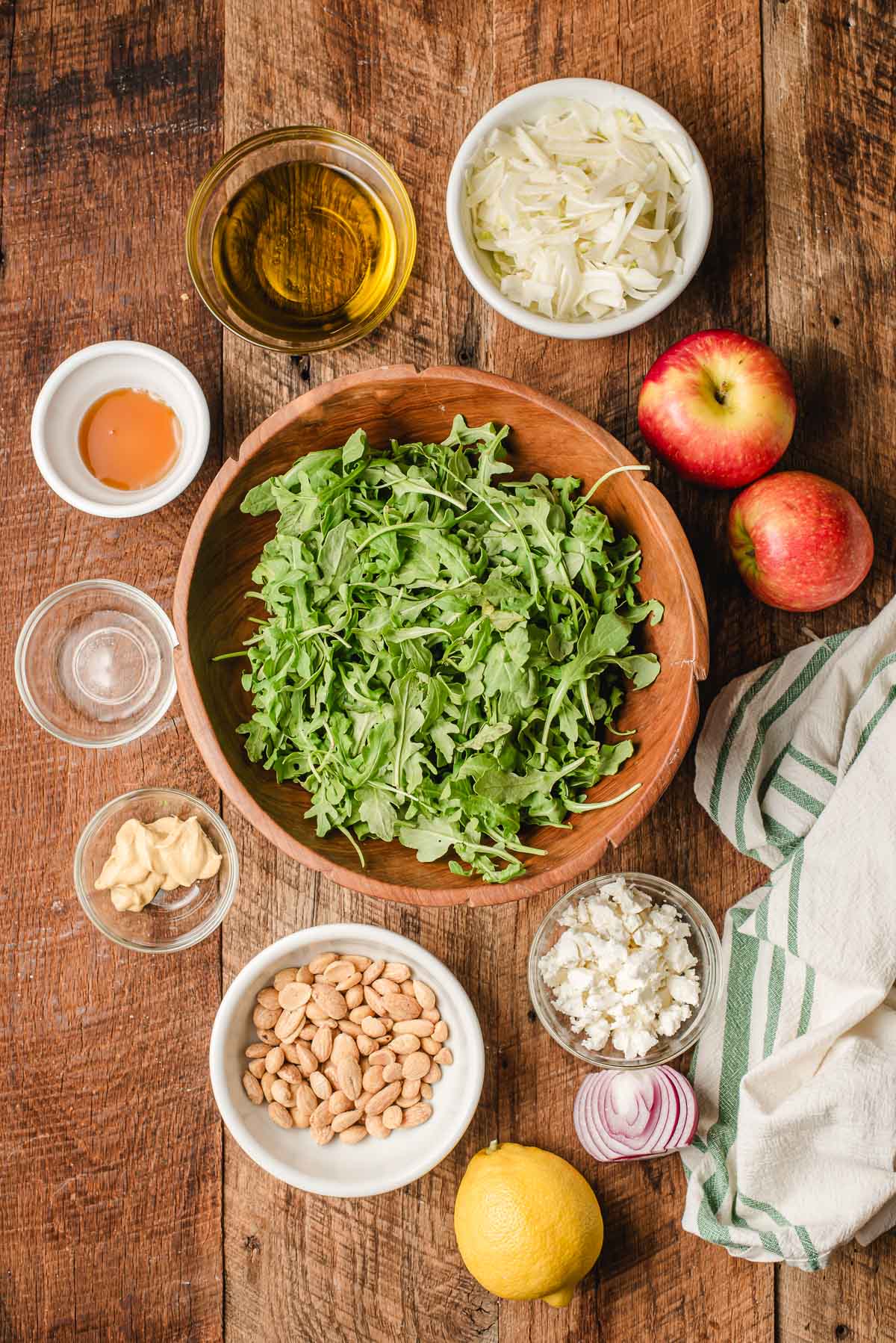 Arugula, marcona almonds, goat cheese, dijon mustard, olive oil, lemon juice, apples, fennel, rice vinegar, and red onion on a wooden background.