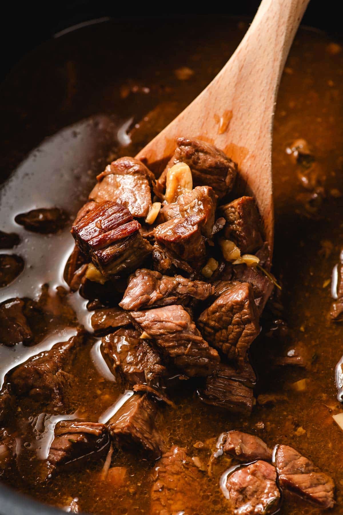 Wooden spoon scooping garlic butter steak bites out of a slow cooker.