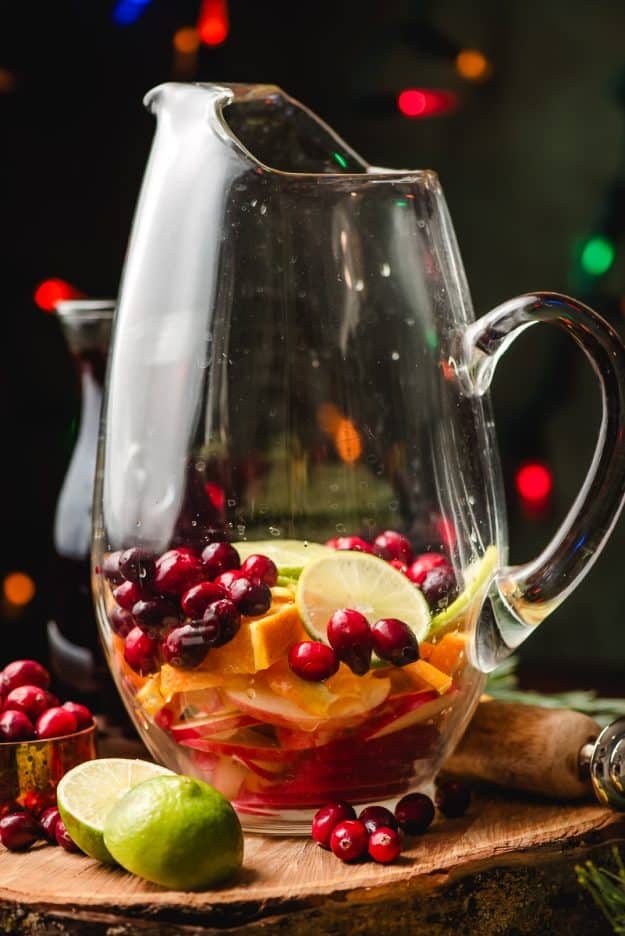Glass pitcher filled with sliced apples, oranges, limes, and cranberries.