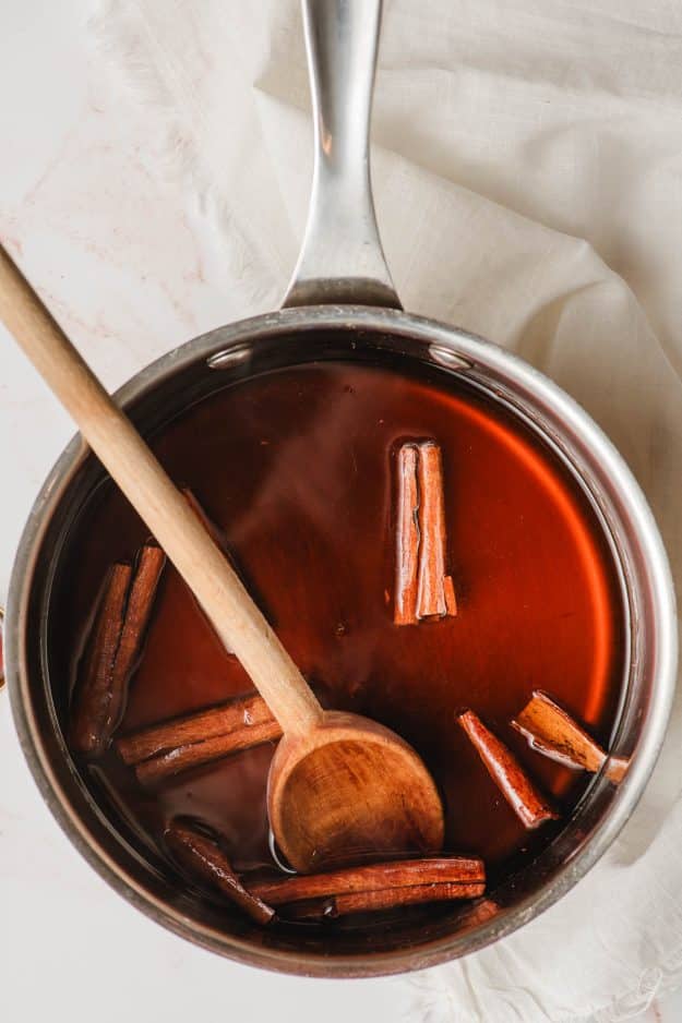 Cinnamon syrup with cinnamon sticks in a saucepan being stirred by a wooden spoon.