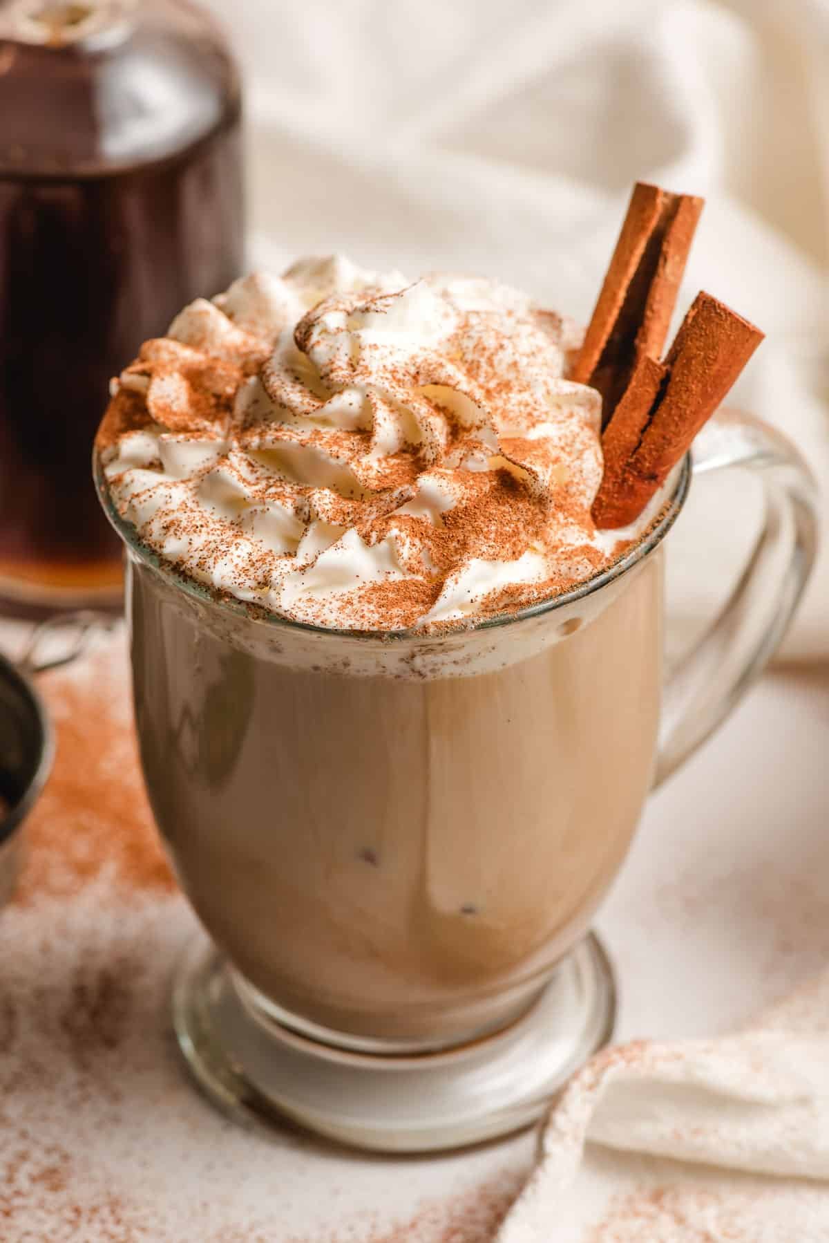 Glass mug filled with a cinnamon spiced latte and topped with whipped cream, cinnamon sticks, and ground cinnamon.
