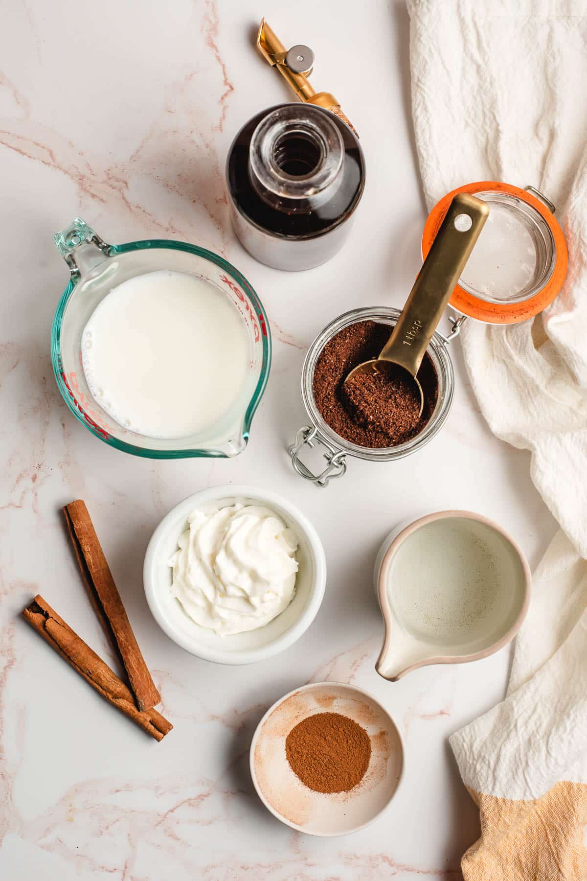 Cinnamon sticks, coffee, water, cinnamon, whipped cream, milk, and cinnamon syrup shown in prep bowls or glass measuring cups.