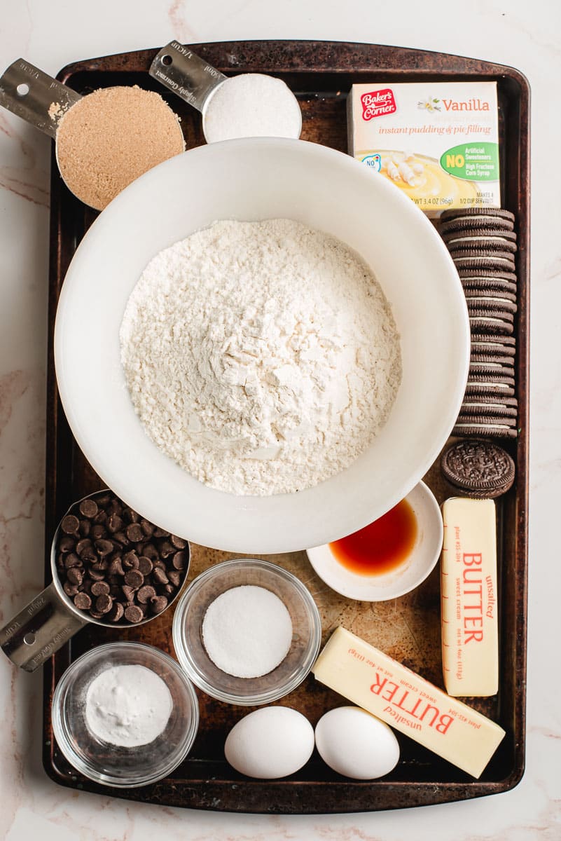 Flour, sugar, brown sugar, eggs, baking soda, salt, vanilla pudding mix, butter, and Oreos shown on a sheet pan.