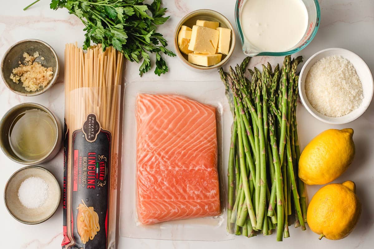 Salmon, asparagus, lemons, Parmesan cheese, heavy cream, butter, parsley, garlic, linguine, and salt and pepper on a white background.