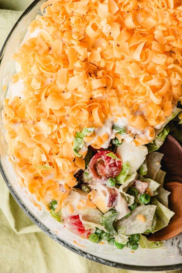 Wooden spoon lifting a serving of 7 layer salad showing creamy layers of lettuce, bacon, and eggs