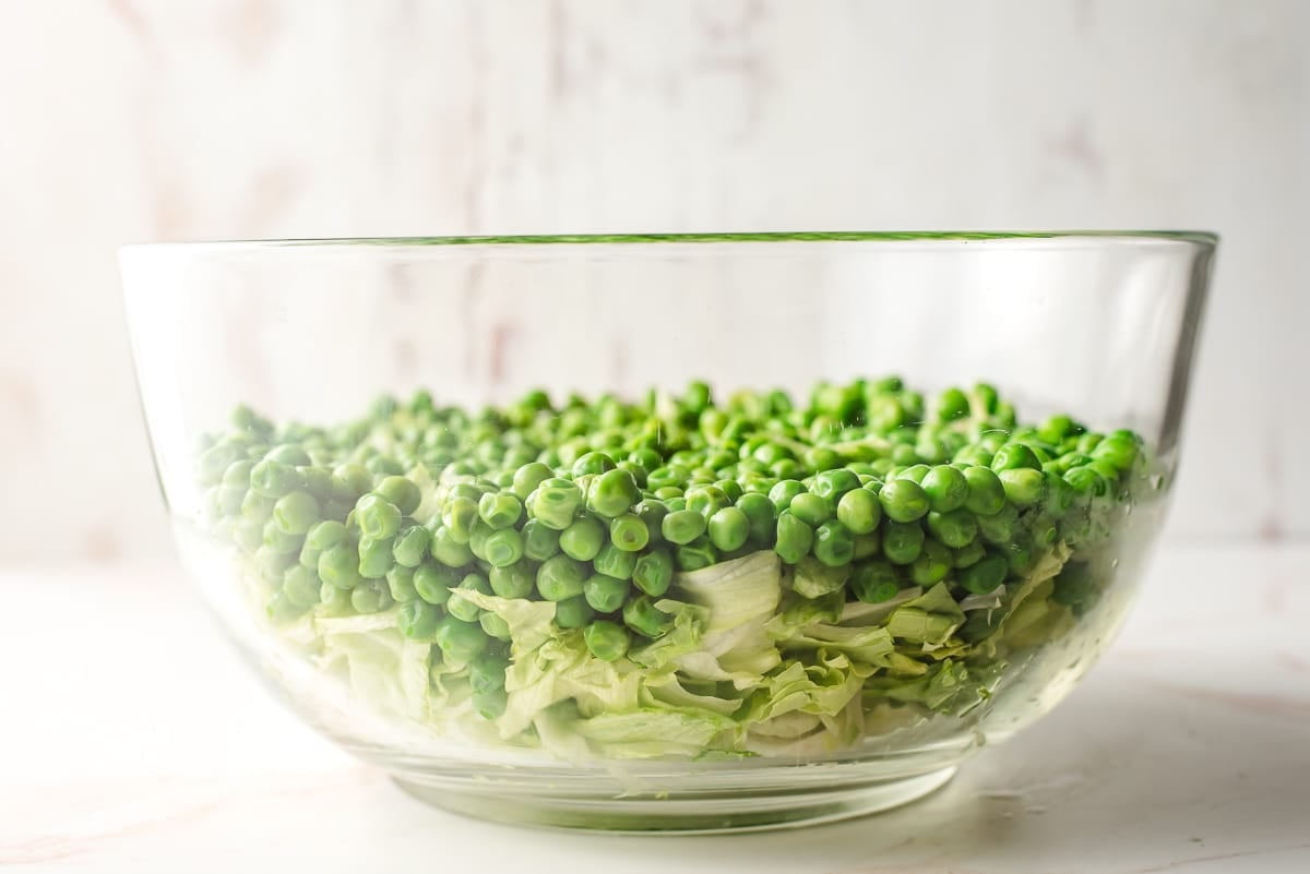 Iceberg lettuce and peas layered in a glass mixing bowl.