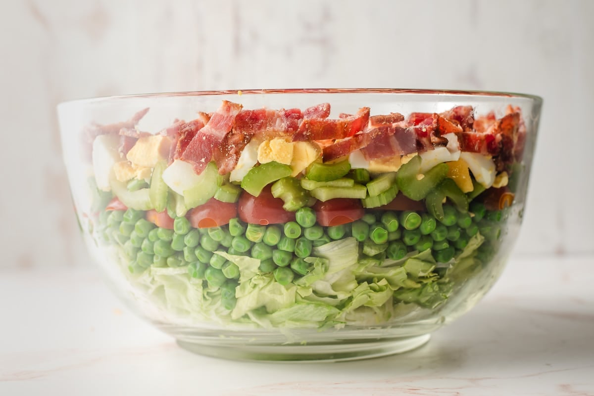 Large glass mixing bowl with lettuce, peas, tomatoes, celery, hardboiled eggs, and bacon.