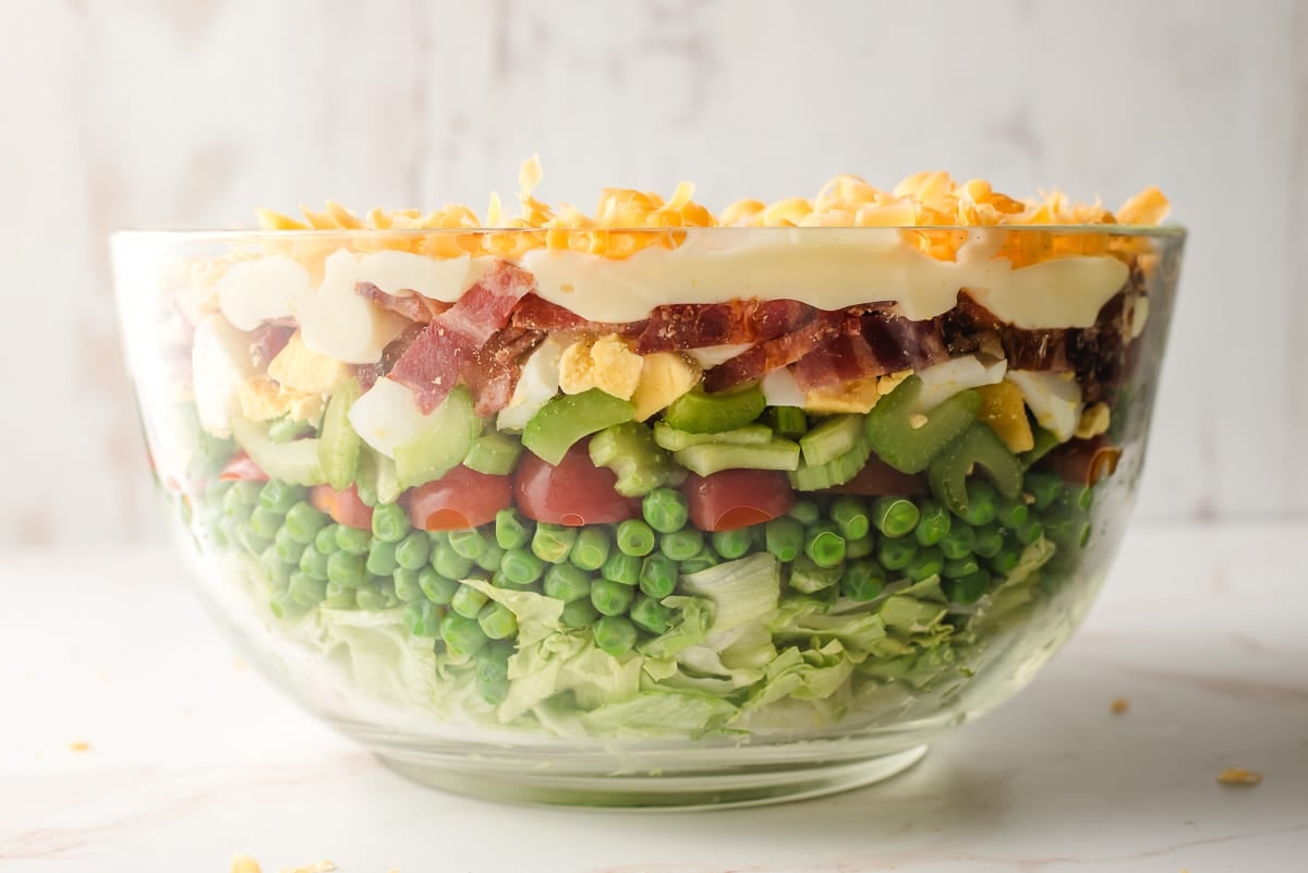 Salad layered with lettuce, peas, tomatoes, celery, hard boiled eggs, bacon, mayonnaise dressing, and cheddar cheese.