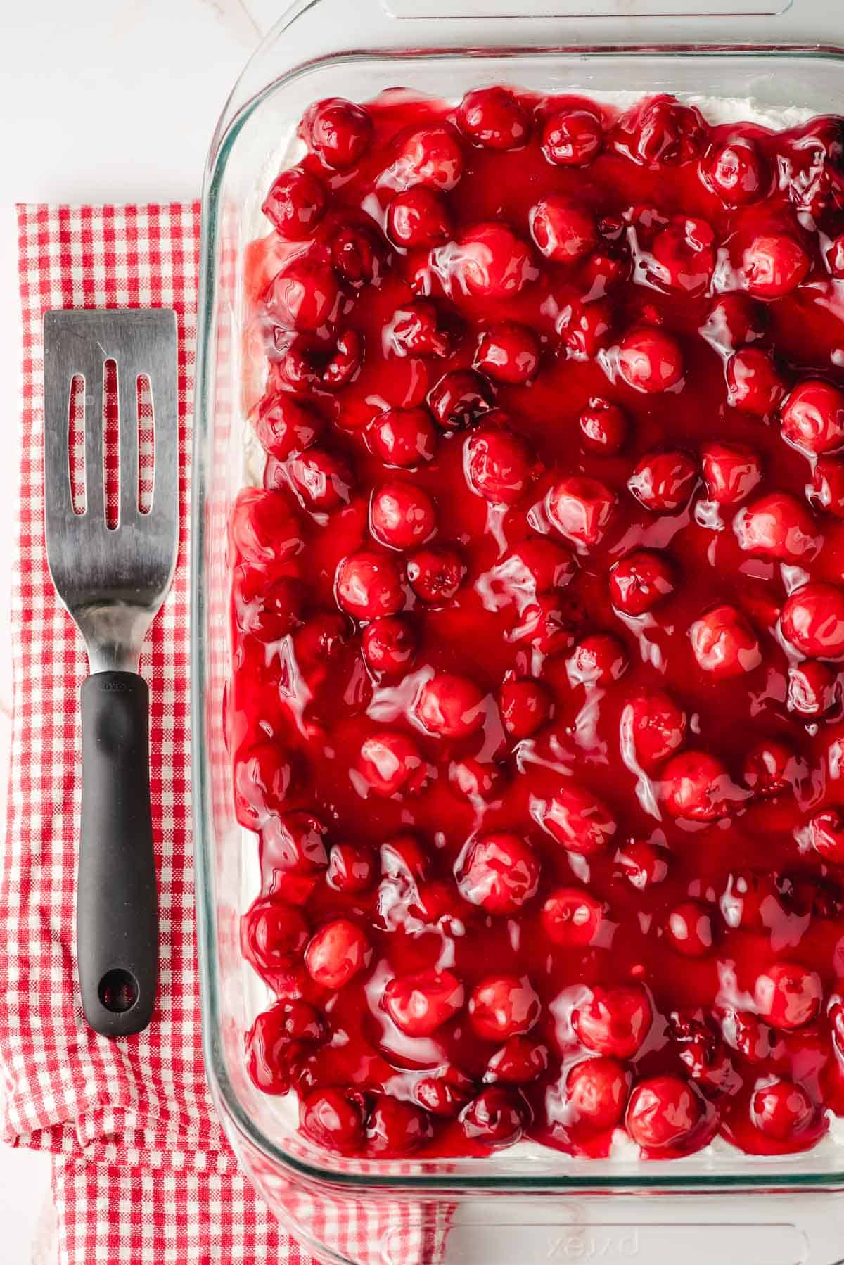 Cherry delight in a glass dish with a red checked tea towel and metal spatula beside it.
