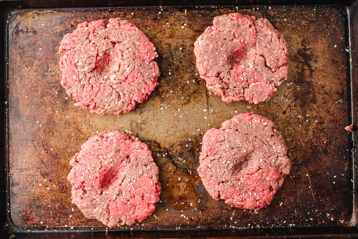 Four raw burger patties on a baking sheet with salt and pepper.