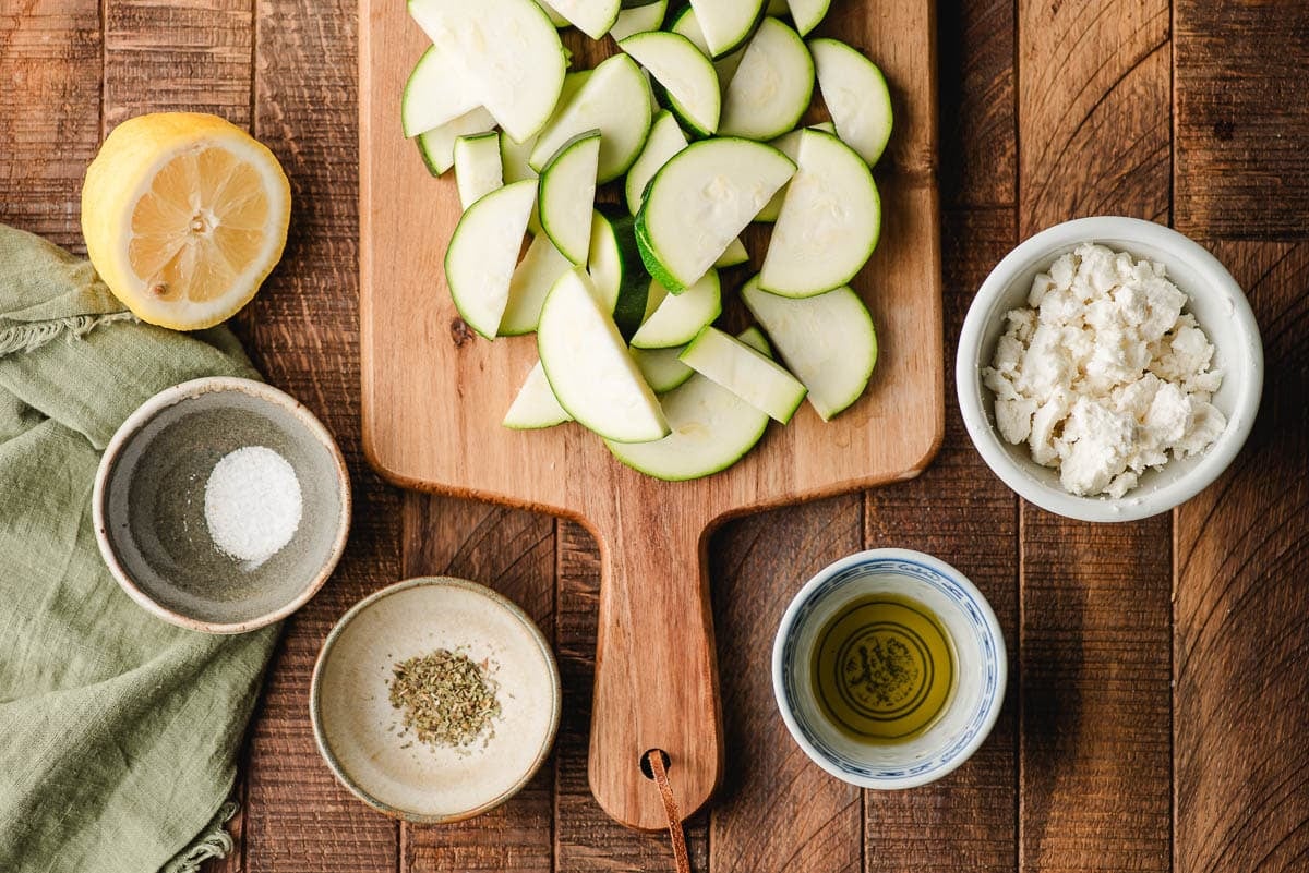 Sliced zucchini on a cutting board, surrounded by bowls of goat cheese, olive oil, seasonings, and a lemon.