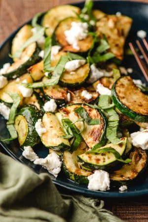 Zucchini saute with goat cheese and fresh basil on a navy blue plate.