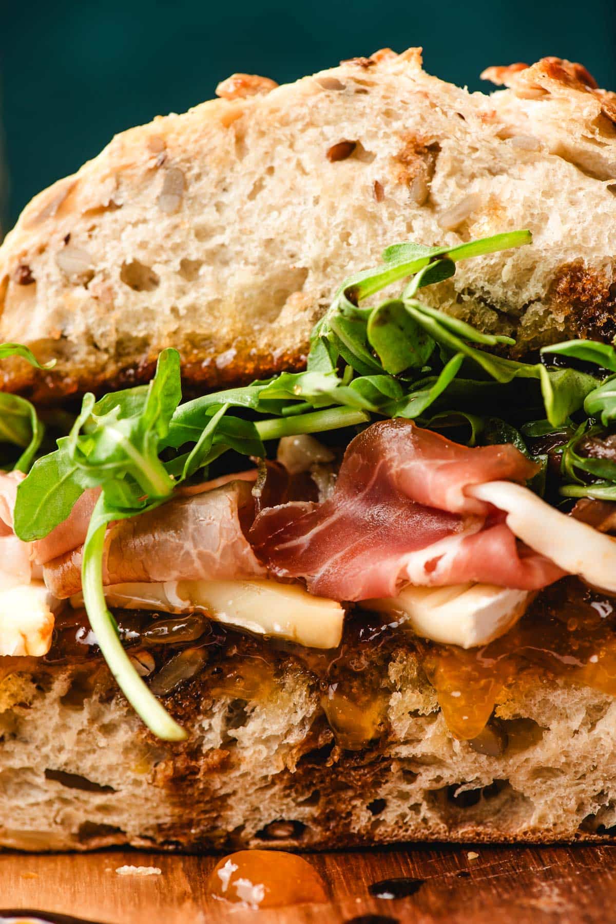 Tight, close up image of sandwich stacked with apricot jam, brie cheese, prosciutto, arugula, and balsamic vinegar.