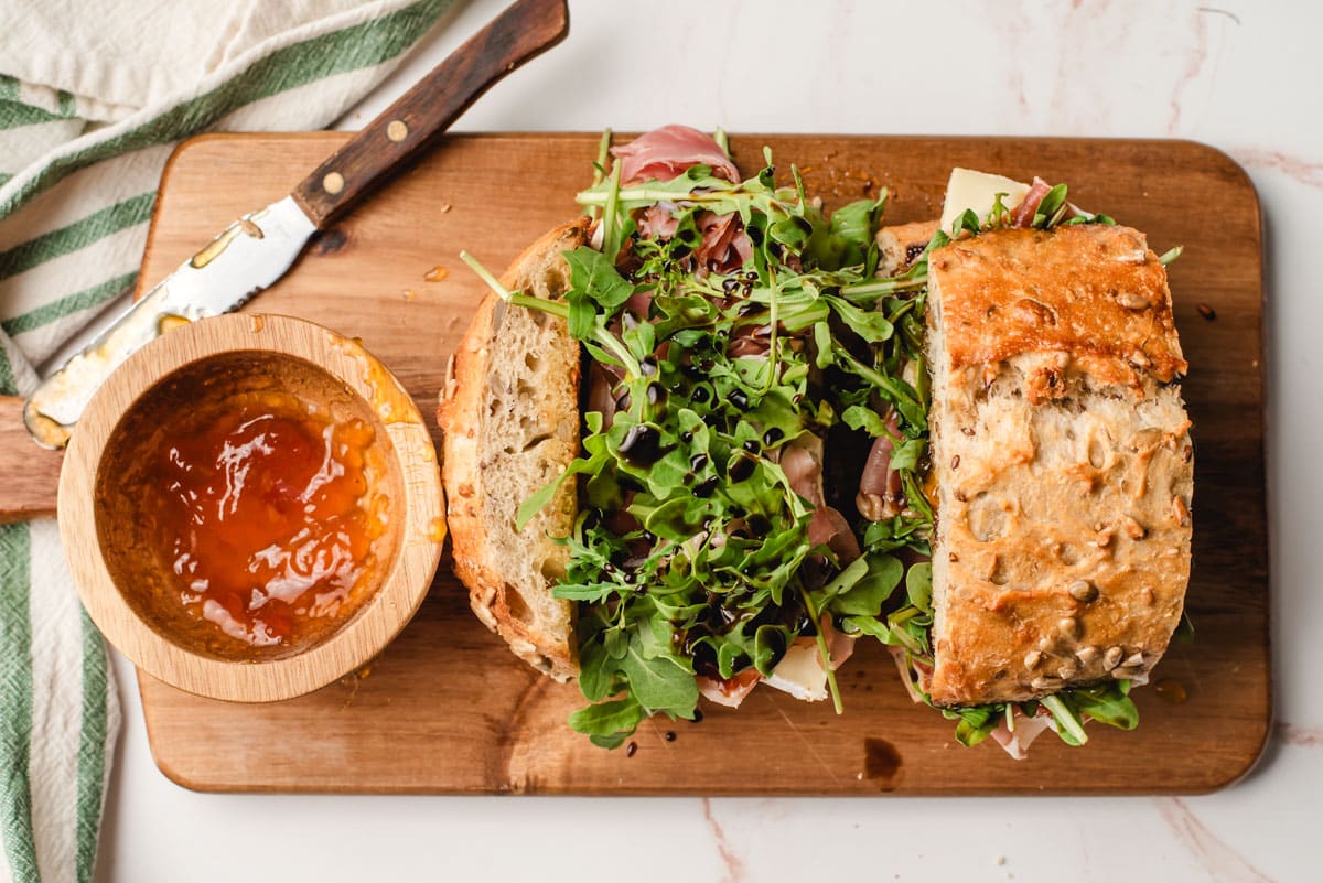 Sandwich shown open face with brie, prosciutto, arugula, and drizzle of balsamic vinegar.