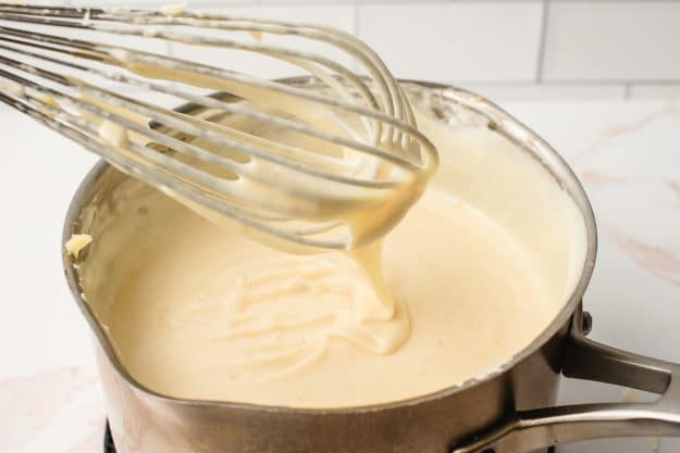 Whisk showing the creamy texture of a roux-based cheese sauce.